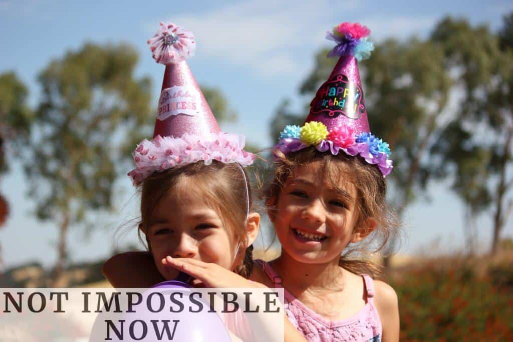 girl in left blowing purple balloon beside girl wearing pink birthday hat
