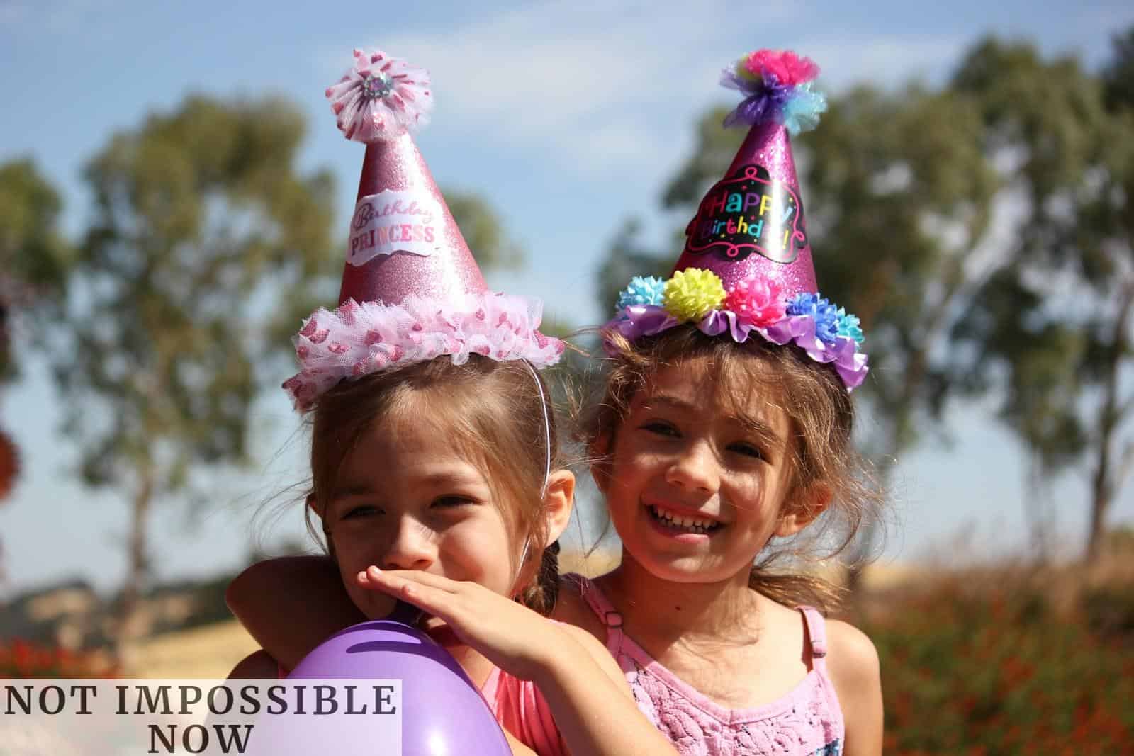 girl in left blowing purple balloon beside girl wearing pink birthday hat